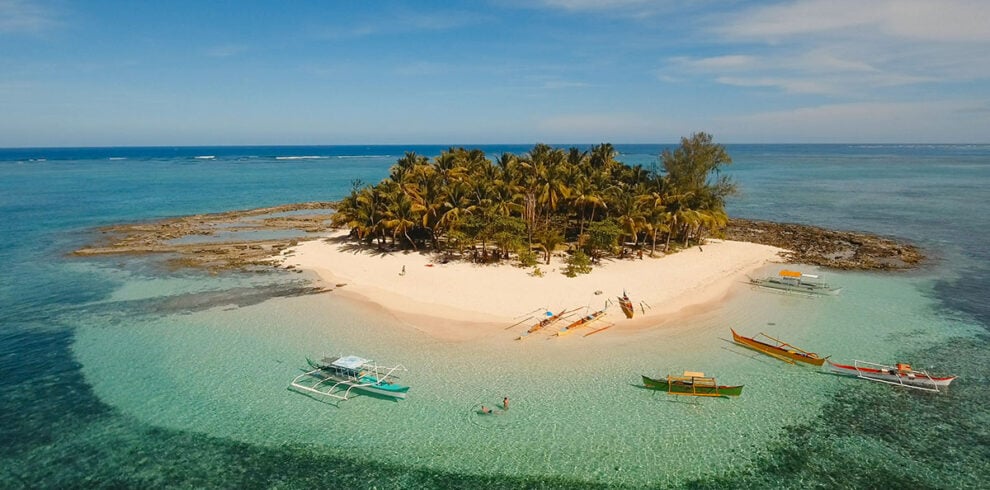 Luchtfoto van Guyam Island, een tropisch eiland bij Siargao, met een hagelwit zandstrand, wuivende palmbomen, helder turquoise water en een omringende koraalrif.