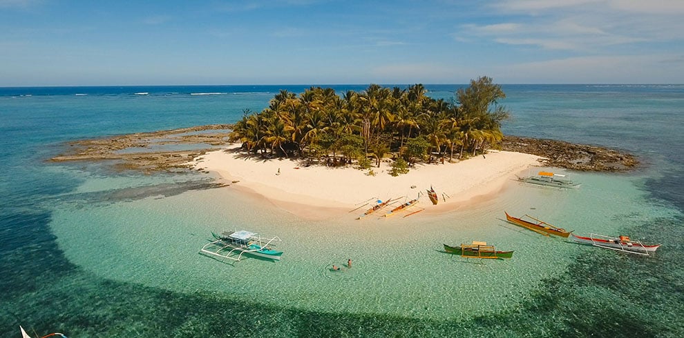 Luchtfoto van Guyam Island, een tropisch eiland bij Siargao, met een hagelwit zandstrand, wuivende palmbomen, helder turquoise water en een omringende koraalrif.