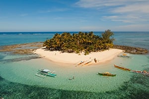 Luchtfoto van Guyam Island, een tropisch eiland bij Siargao, met een hagelwit zandstrand, wuivende palmbomen, helder turquoise water en een omringende koraalrif.